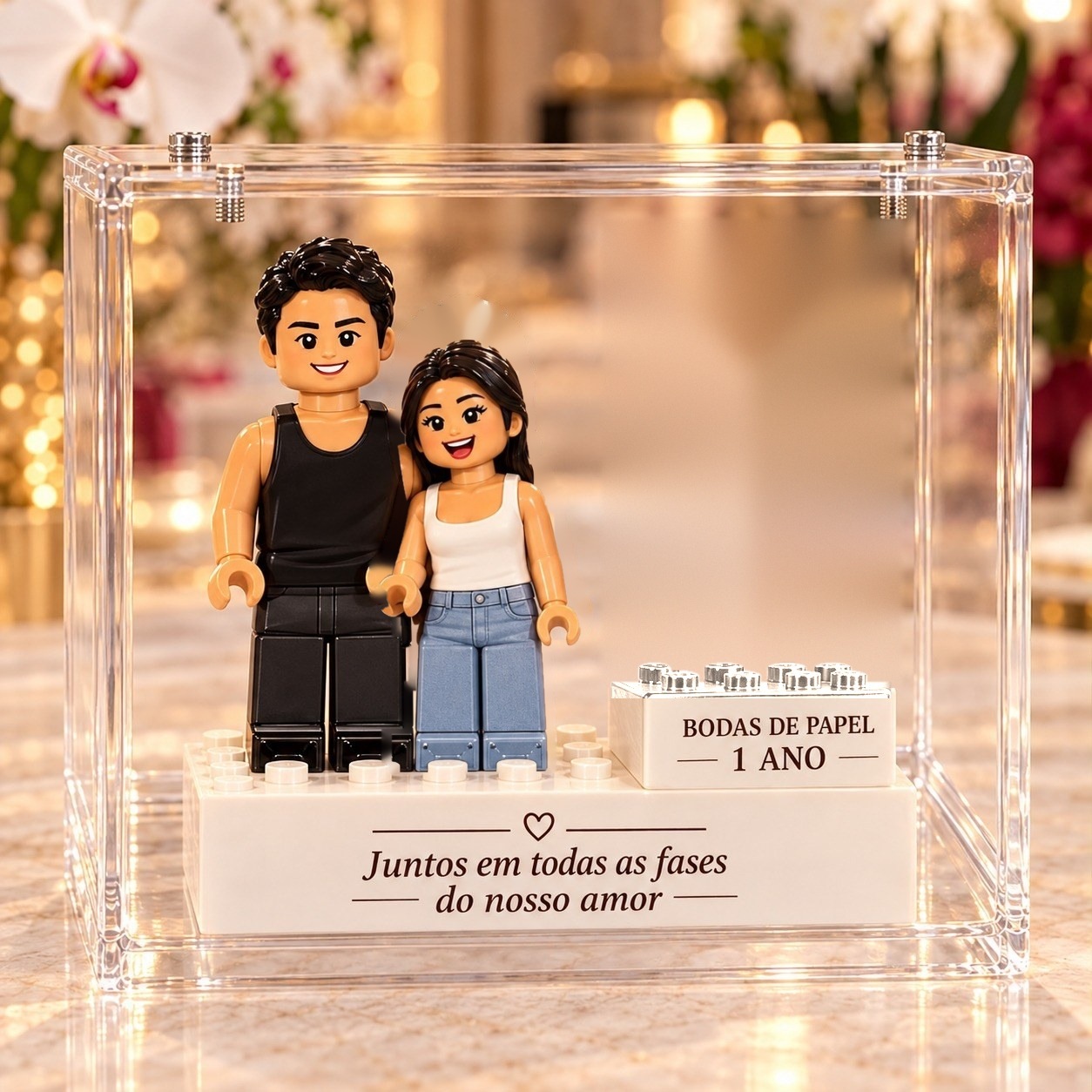 Casal em versão de blocos de montar, ele de regata preta e ela de regata branca e jeans, ao lado do bloco das bodas de papel, numa vitrine de acrílico com a frase Juntos em todas as fases do nosso amor